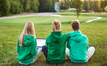 3 opiskelijaa istuu nurmikolla, yllään vihreät hupparit. Kuva on otettu takaapäin. Kuva on luotu Adobe firefly-tekoälytyökalulla.